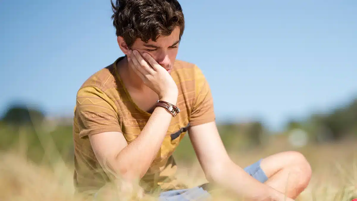 Teen boy sitting alone in a field. Supportive societal and institutional cultures can promote teen mental health, recovery, and well-being.